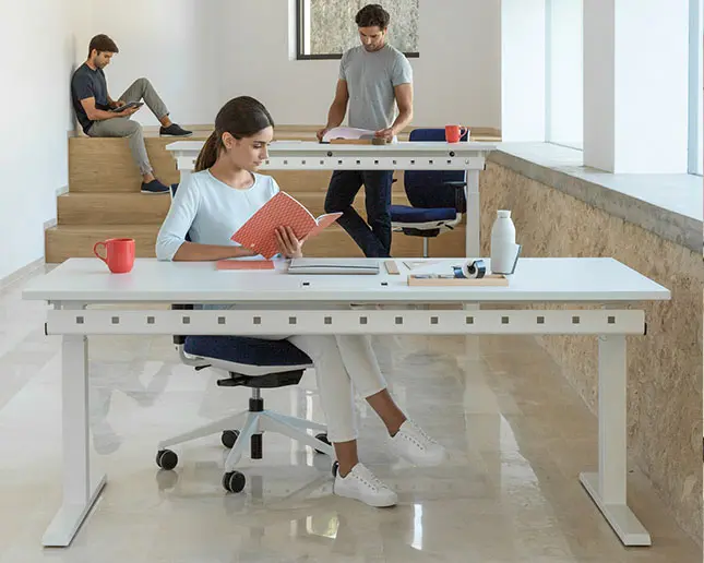 Mesa elevable eléctrica regulable en altura para mejorar la salud laboral Jóvenes estudiando y trabajando en mesa de oficina Actiu Mobility Step con estructura elevable regulable en altura